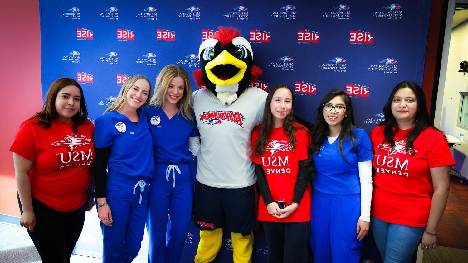 六名医疗保健专业的女学生在丹佛州立大学的蓝色媒体背景前与Rowdy合影